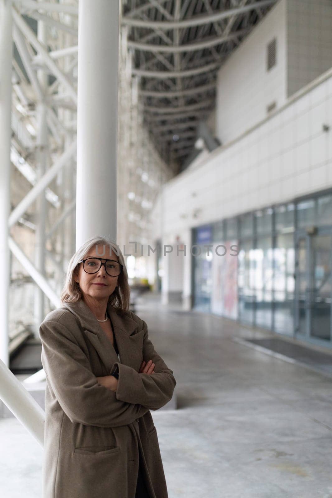 An elderly businesswoman poses confidently in a spacious, modern urban area, arms crossed, reflecting her experience and professional outlook in a contemporary setting.