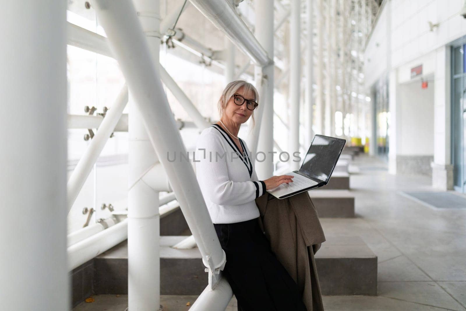An elderly businesswoman is focused on her laptop, sitting comfortably in a modern office environment.