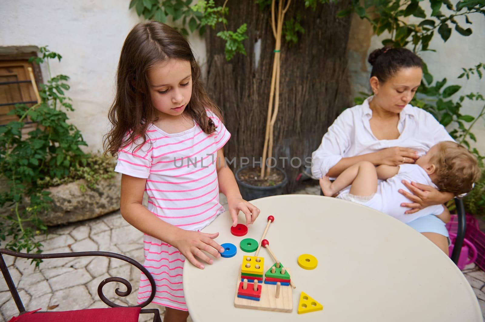 A mother breastfeeds her baby while her young daughter plays with colorful educational toys on a table in an outdoor setting. The image captures a warm family moment.