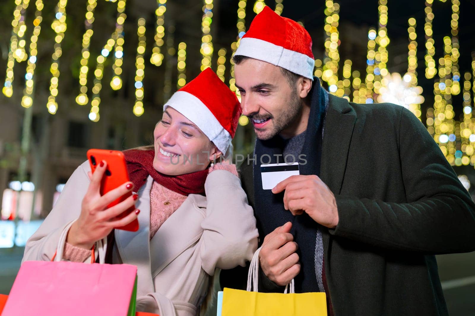 A cheerful couple shops online using a smartphone and credit card under glowing holiday lights. The woman holds shopping bags while they enjoy the festive atmosphere in an outdoor urban setting