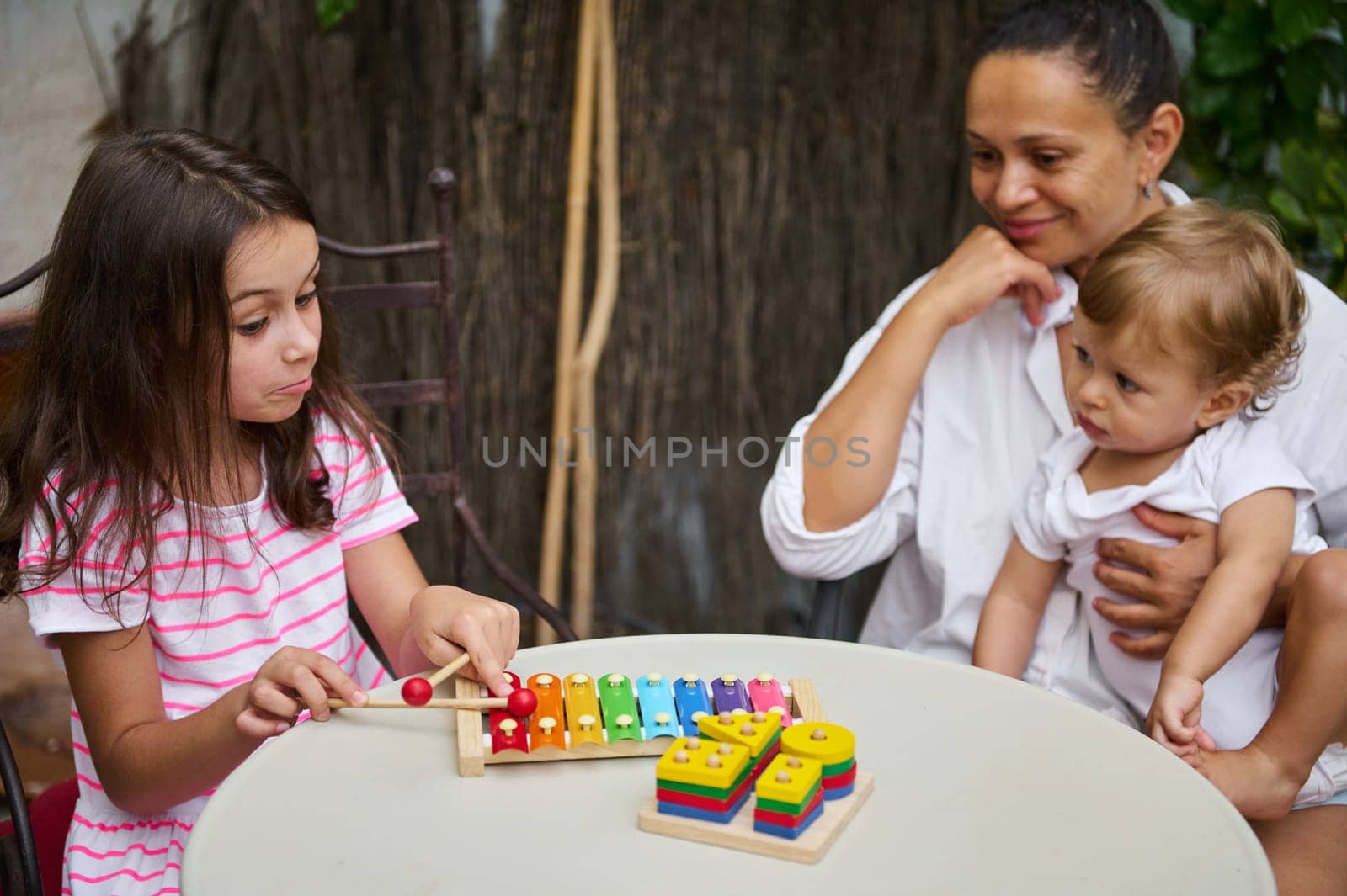A girl plays a colorful xylophone while her mother and baby sibling watch attentively, sharing a joyful moment.