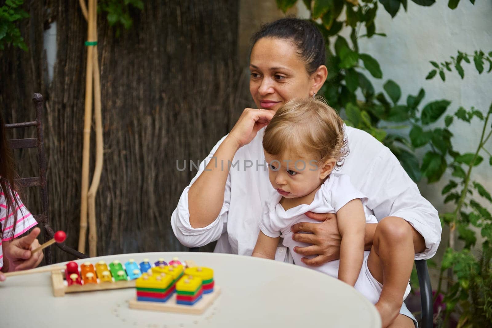 A mother lovingly holds her child while watching colorful toys on a table outside. The scene exudes warmth, togetherness, and maternal bonding in a natural garden setting.