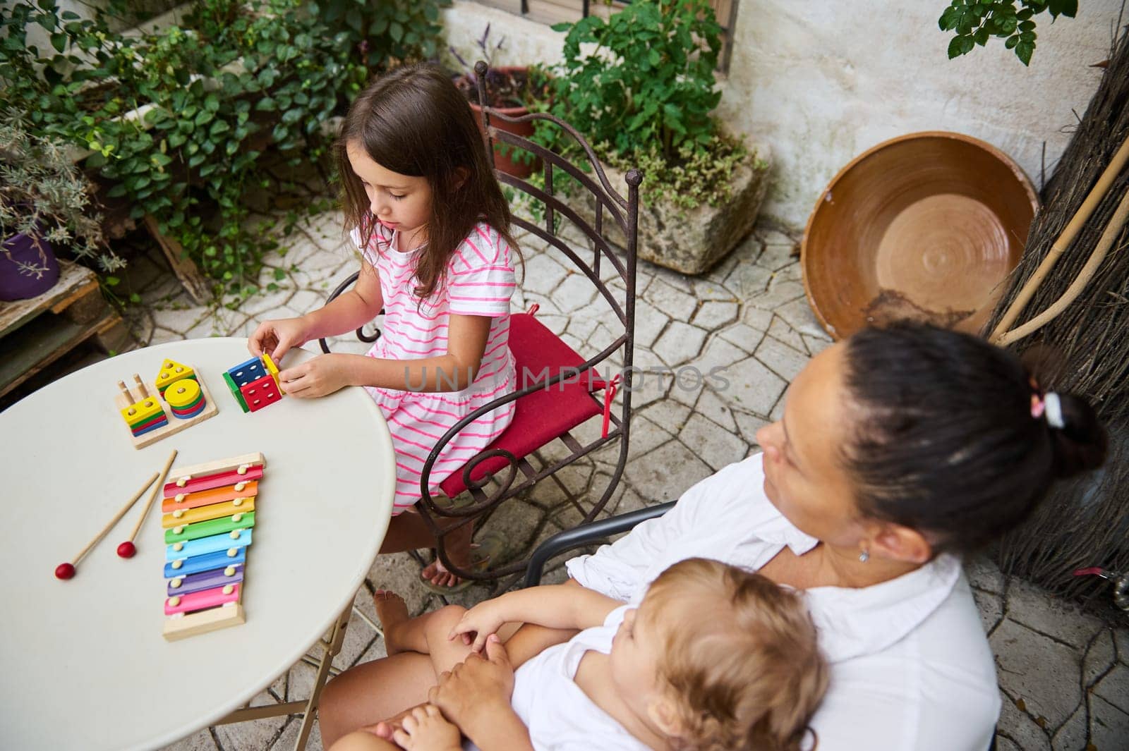 A young girl plays with colorful musical toys at a garden table, while a mother and baby watch. The scene exudes happiness and family bonding, set in a vibrant and cozy outdoor space.