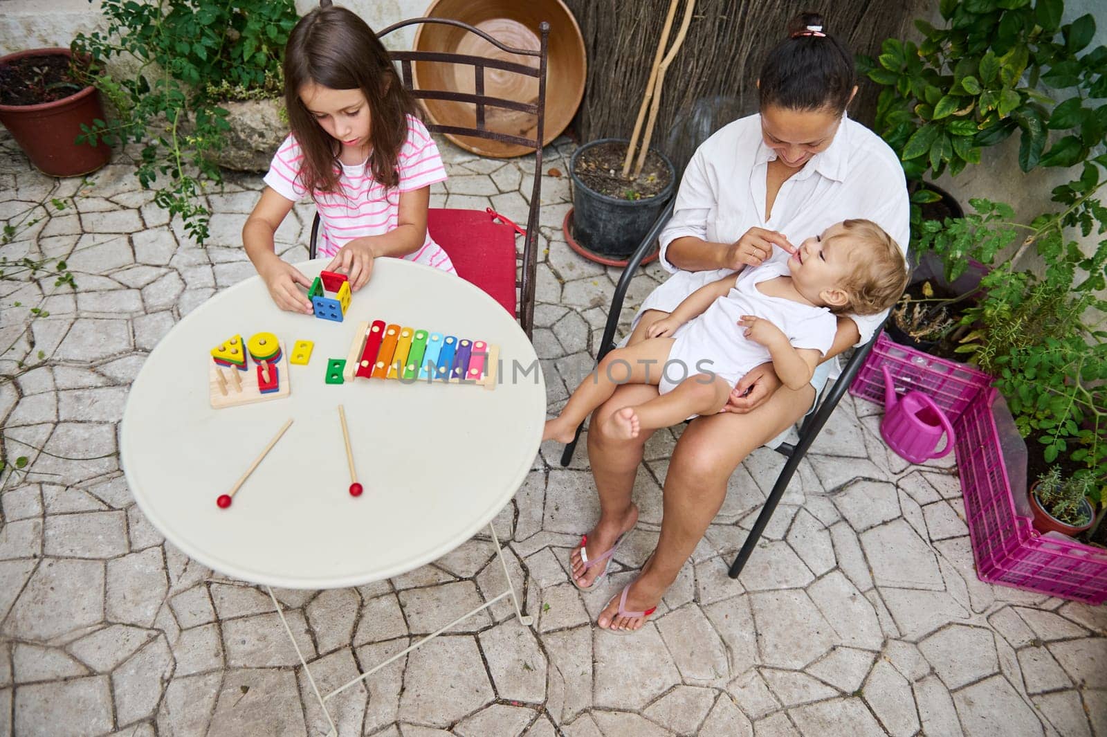 A mother lovingly holds her baby while her daughter plays with colorful educational toys in a garden setting.