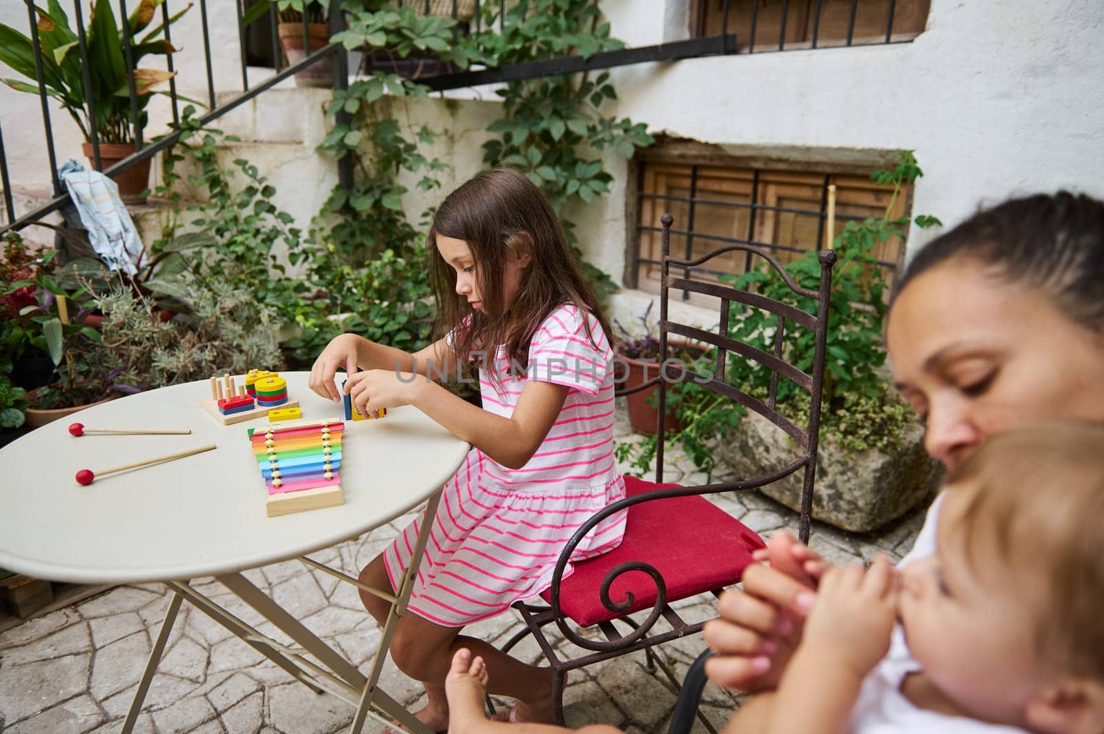 Young girl enjoys playing a colorful xylophone in a cozy garden patio setting with her family nearby.