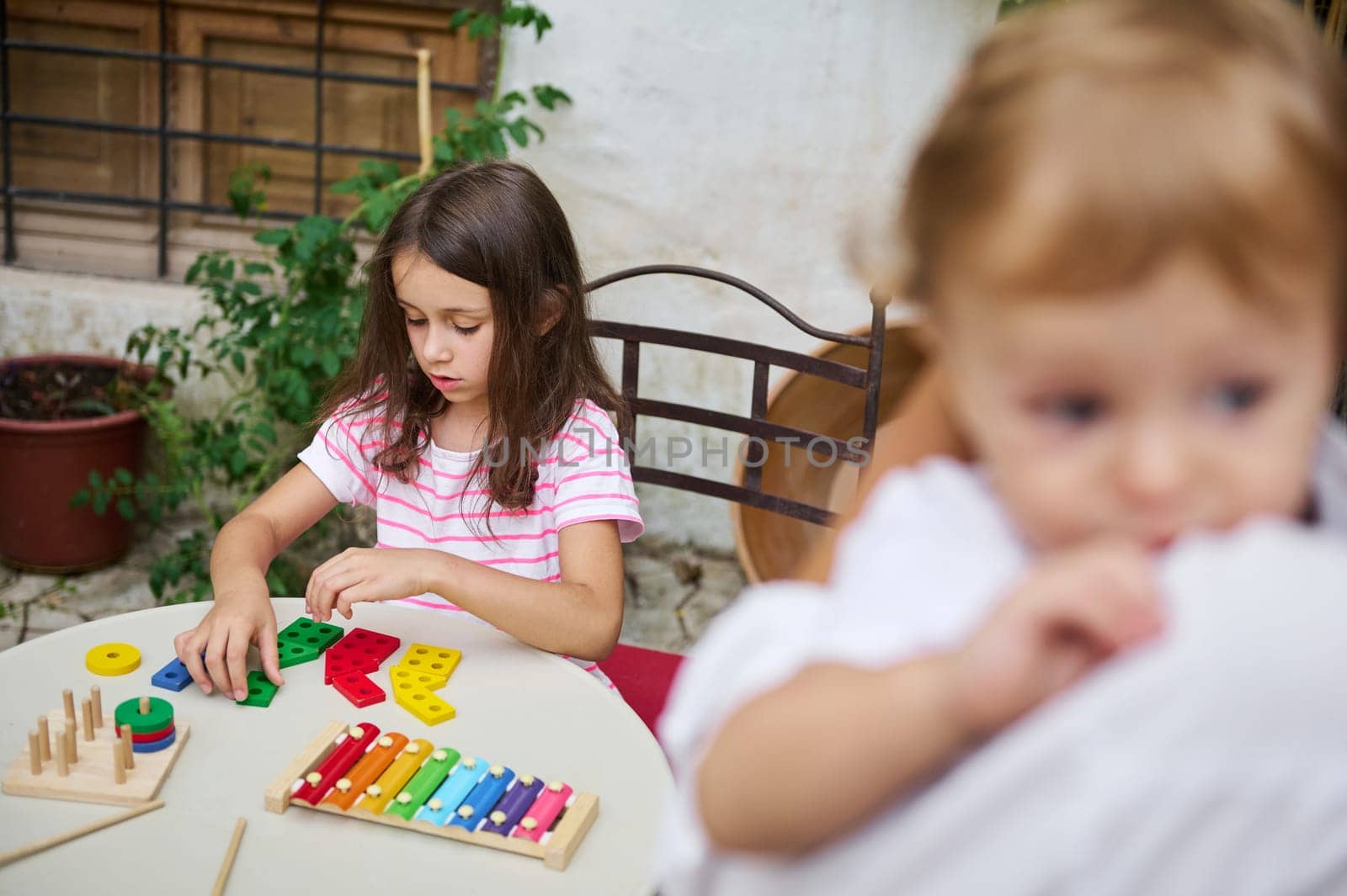 A young girl focuses on playing with colorful educational toys on a table outdoors. A blurred child is visible in the foreground, adding depth to the scene.