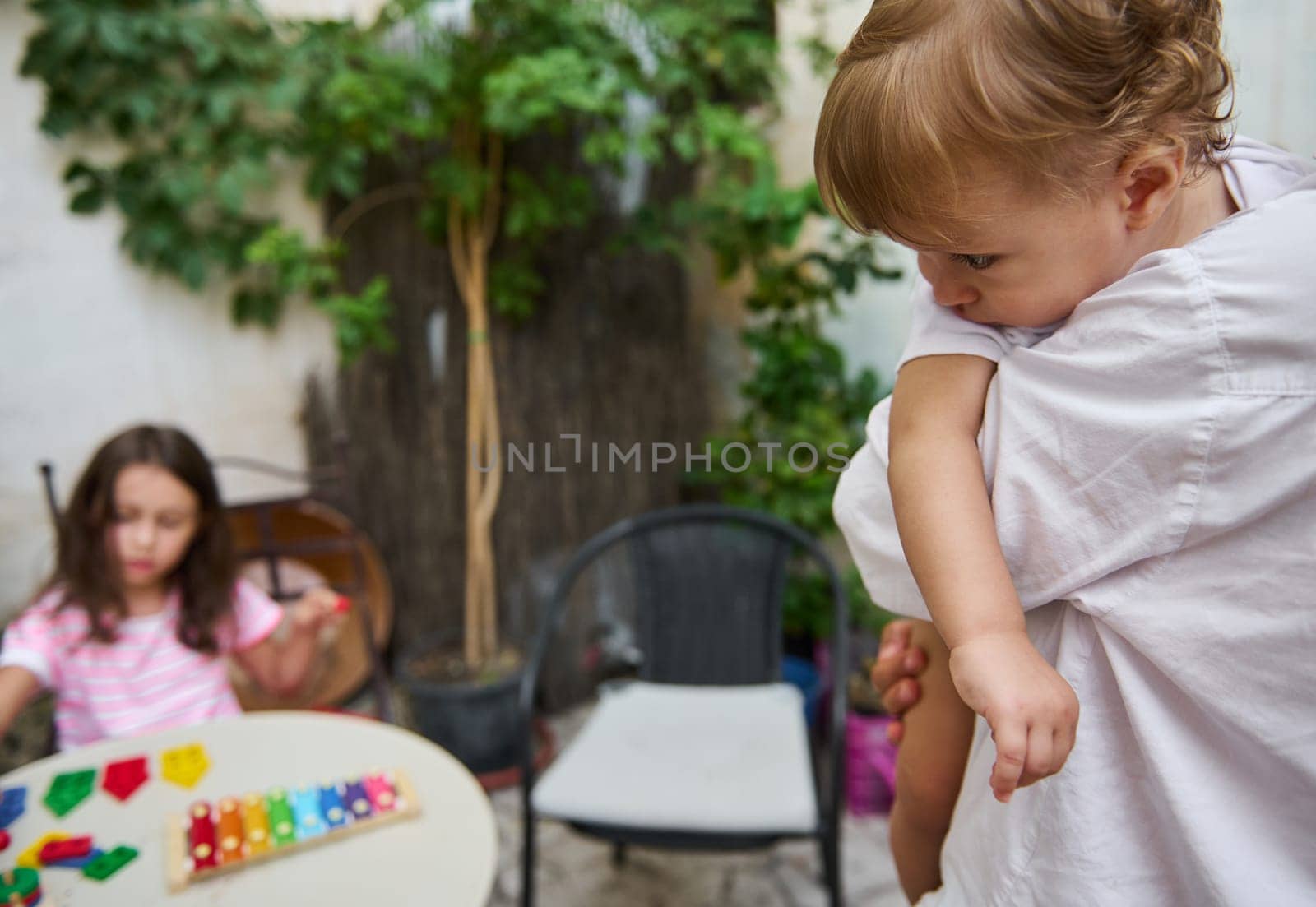 A father gently holds a toddler, capturing a tender moment, while a young girl interacts with vibrant toys on a patio.