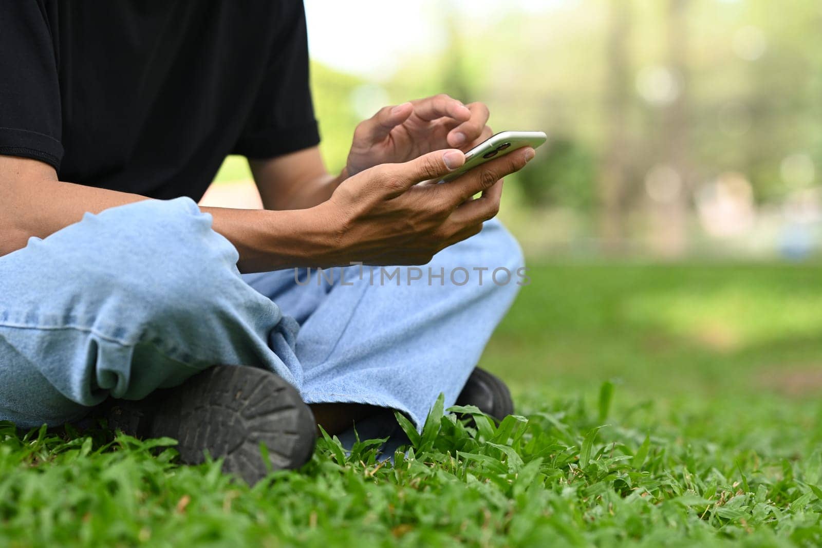 Cropped shot man sitting on the grass in park and checking messages on smartphone.