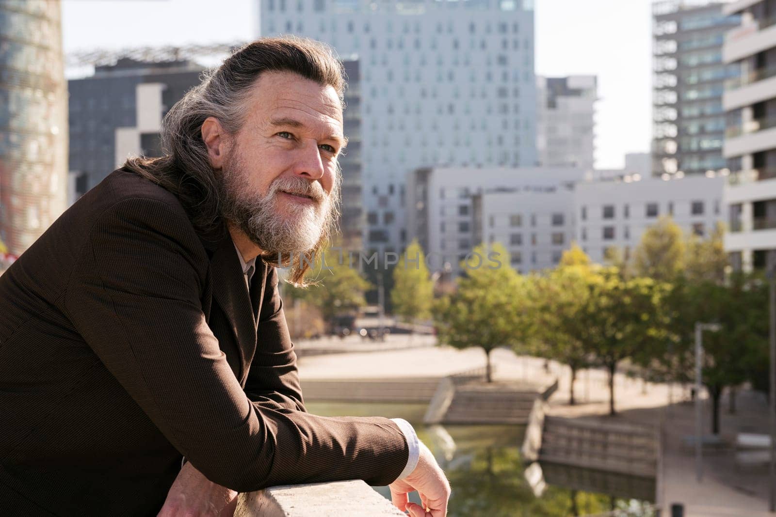 positive middle age male with gray hair and beard in stylish formal outfit looking away pensive while leaning on fence on terrace of city building, concept of business and mature man lifestyle