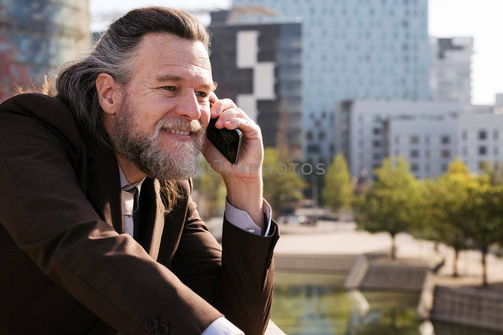 smiling middle age male with beard and gray hair in classy suit discussing business issues on phone conversation against modern city buildings, concept of business and mature man lifestyle