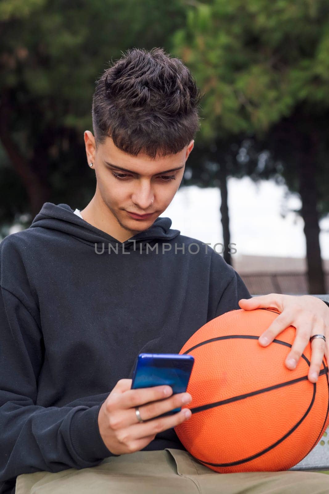 concentrated caucasian teen male using mobile phone sitting with basketball in a bench of a park, concept of youth and technology of communication