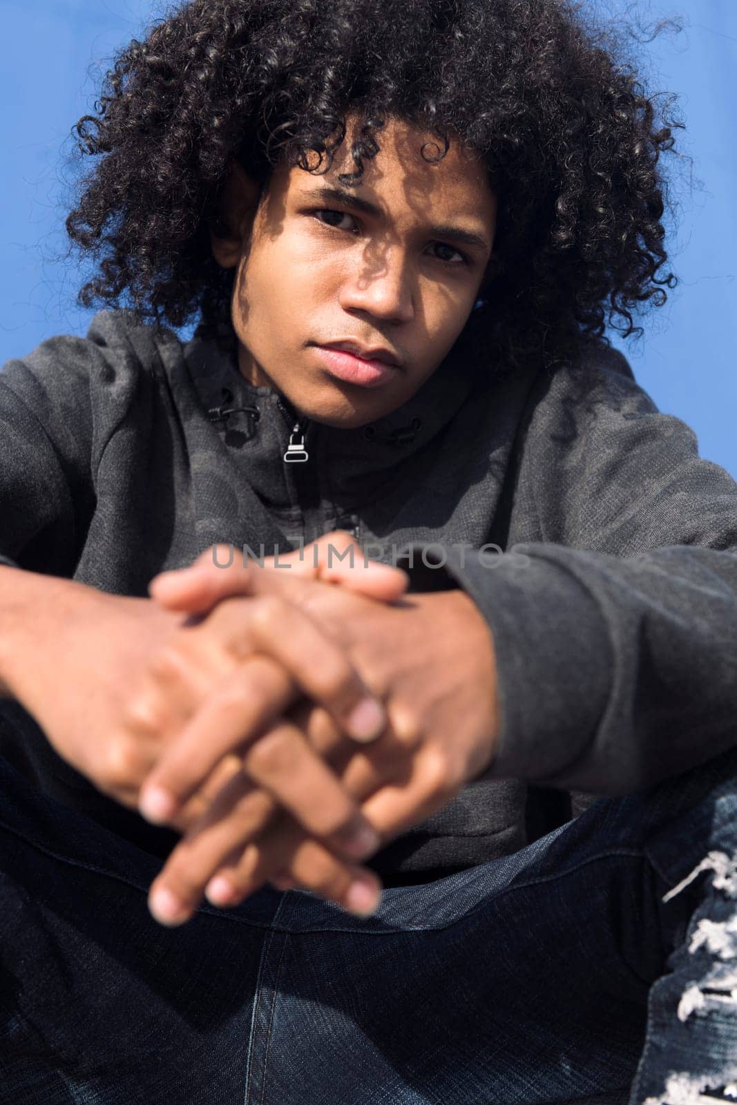 close up portrait of a thoughtful curly haired black male teenager in informal wear looking at camera while sitting against blue background, concept of young lifestyle