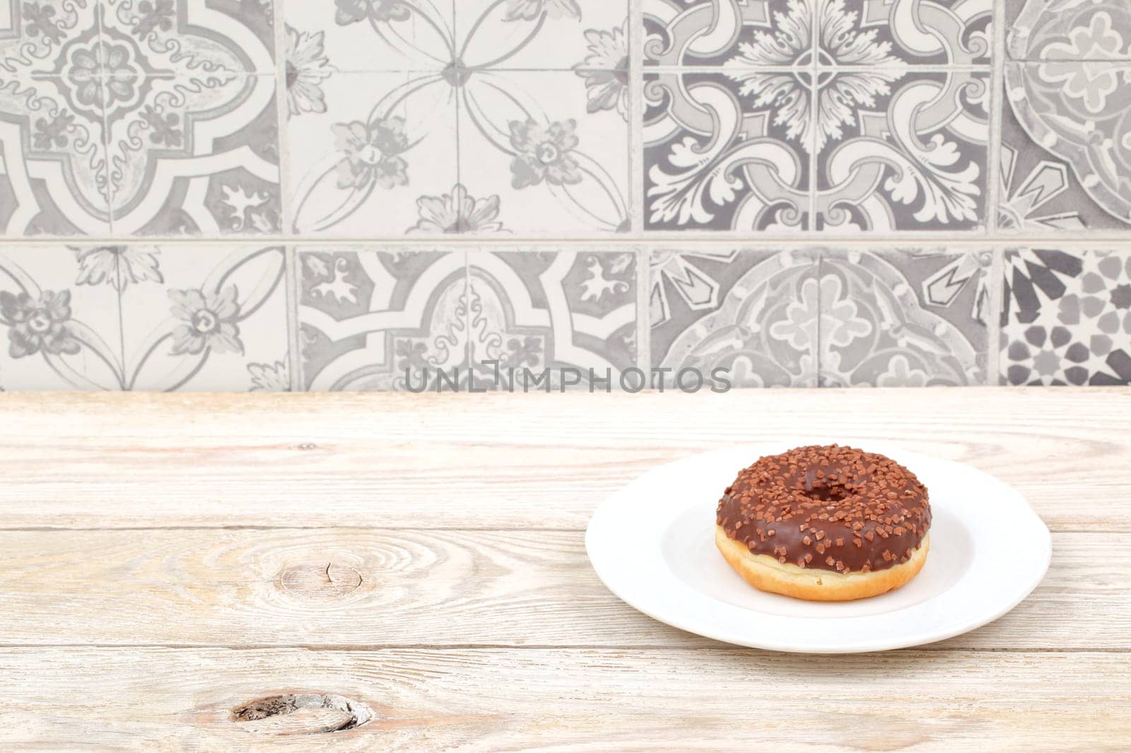 A tempting chocolate donut with colorful sprinkles sits on a white plate against a patterned background