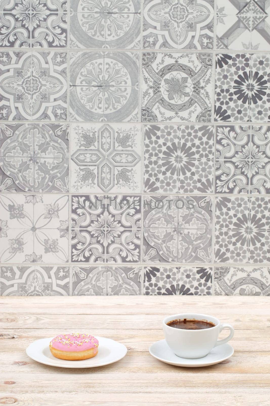 A cozy scene featuring a cup of coffee and a donut on a wooden table against a decorative tile wall.