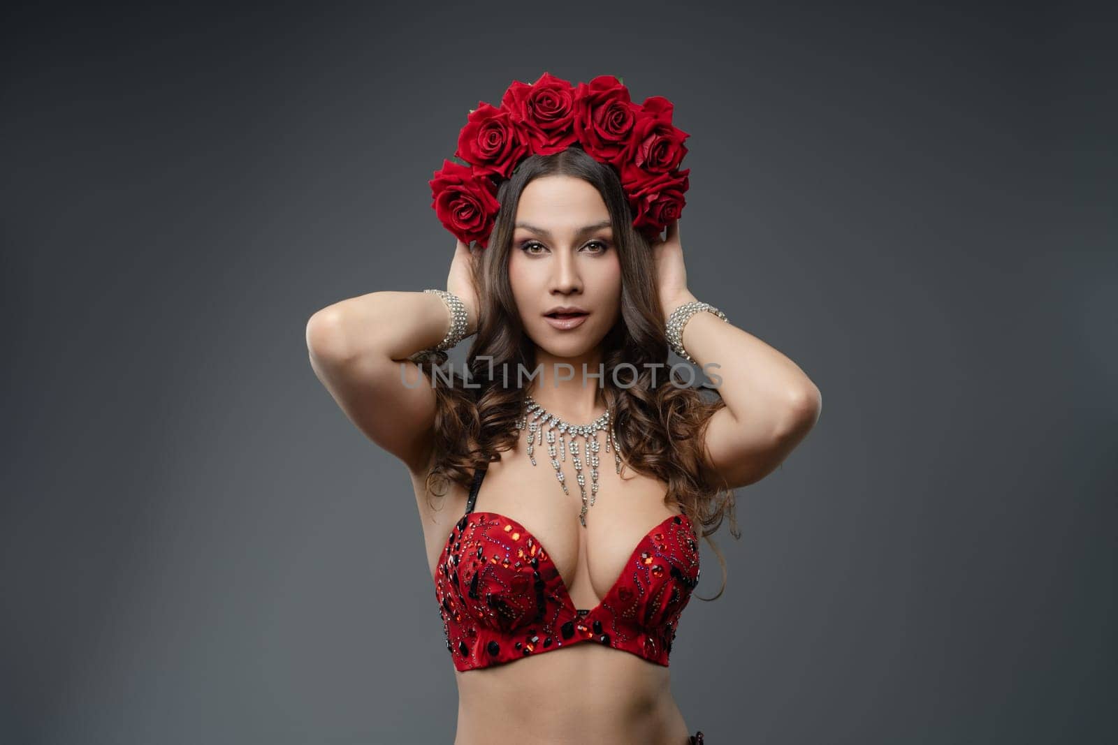 A dancer adorned with a red floral crown and sequined outfit strikes a captivating pose indoors, showcasing her beauty and artistry.