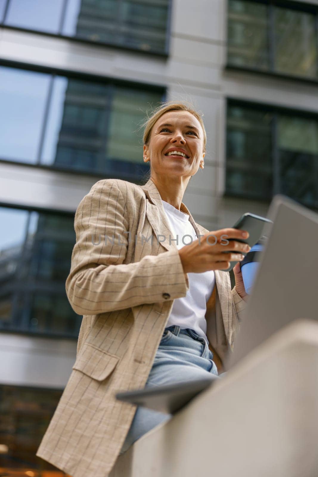 Smiling female entrepreneur use phone while working on laptop in modern office background by Yaroslav_astakhov