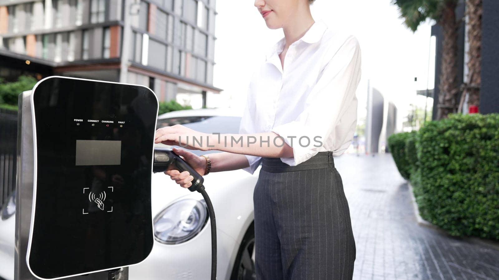 Progressive businesswoman install charger plug from charging station to his electric car before driving around city center. Eco friendly rechargeable car powered by sustainable and clean energy.