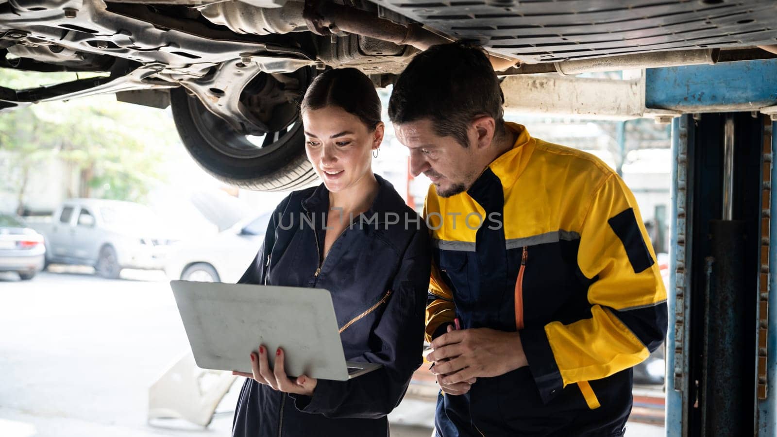 Two vehicle mechanic working together underneath lifted car, conduct car inspection with laptop. Automotive service technician in uniform carefully make diagnostic troubleshooting. Panorama Oxus