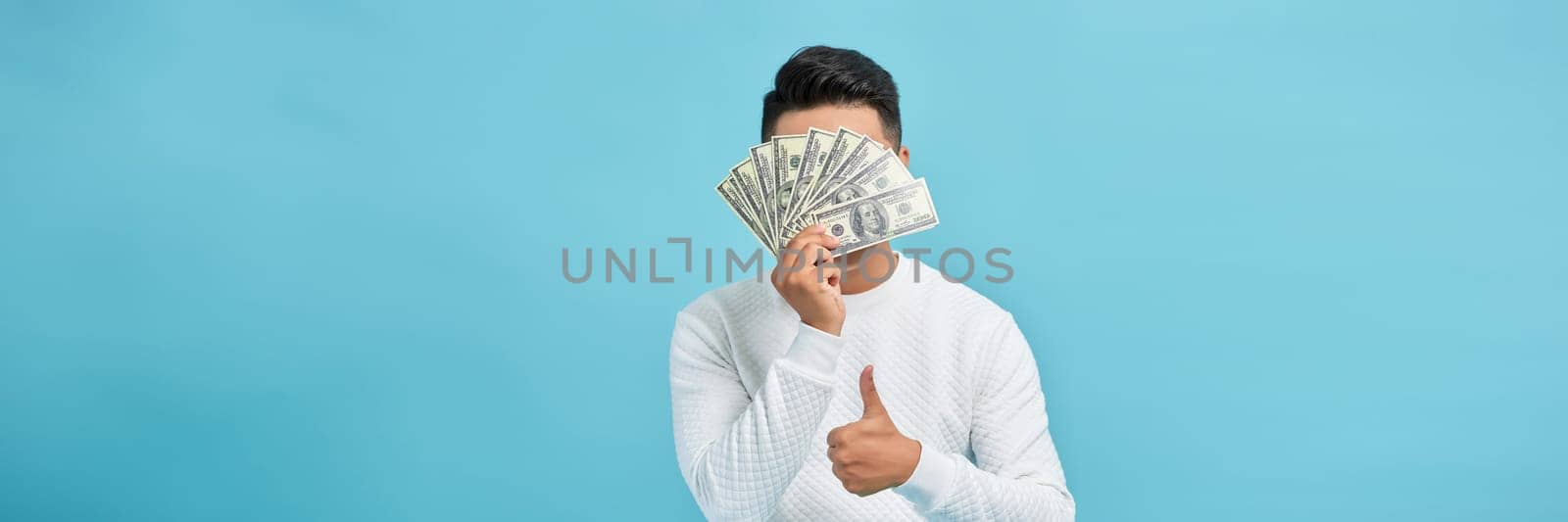Excited young handsome Asian man holding bunch of money banknotes isolated on blue banner