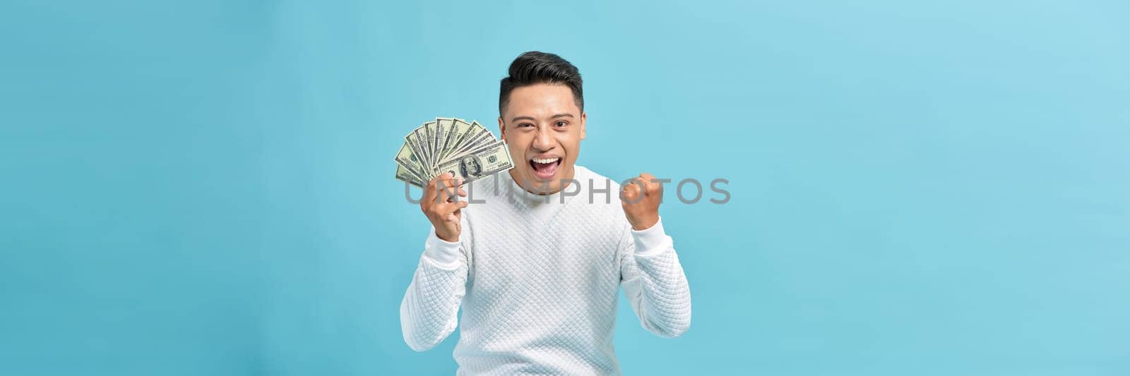 Excited young handsome Asian man holding bunch of money banknotes isolated on blue banner by makidotvn