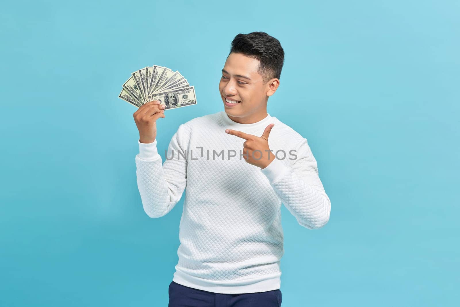 Smiling asian man pointing at fan of money over blue background, copy space