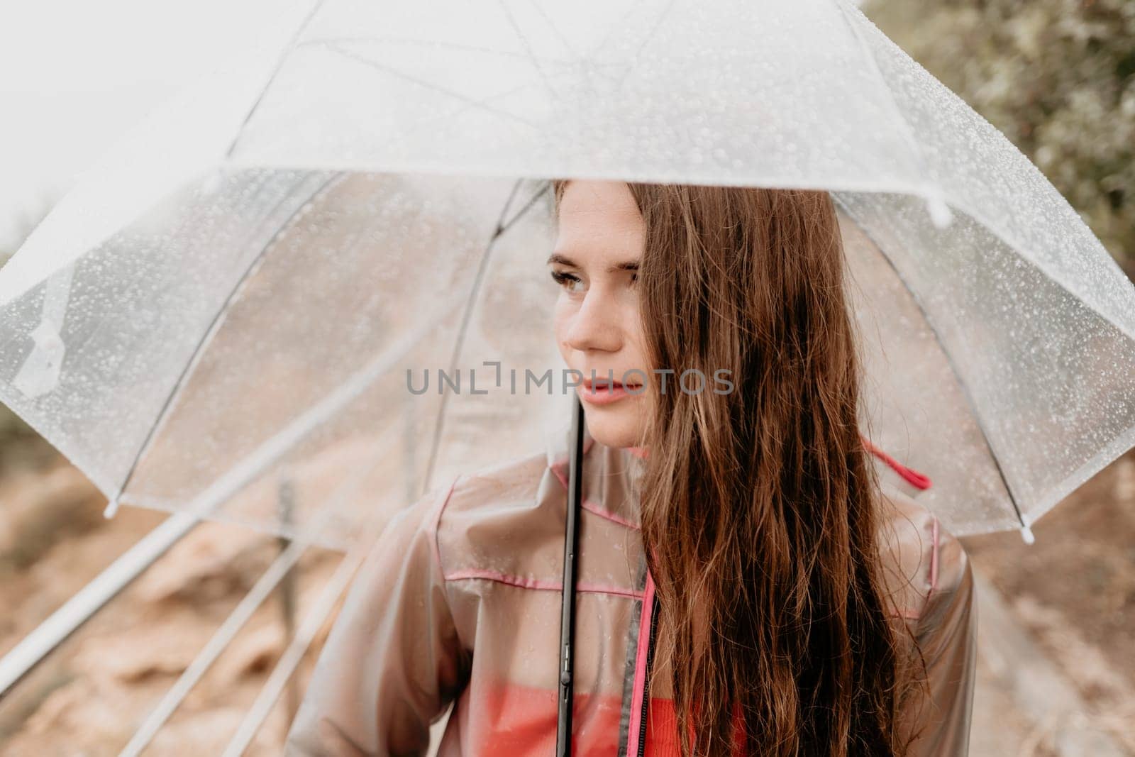 Woman rain umbrella. Happy woman portrait wearing a raincoat with transparent umbrella outdoors on rainy day in park near sea. Girl on the nature on rainy overcast day. by panophotograph