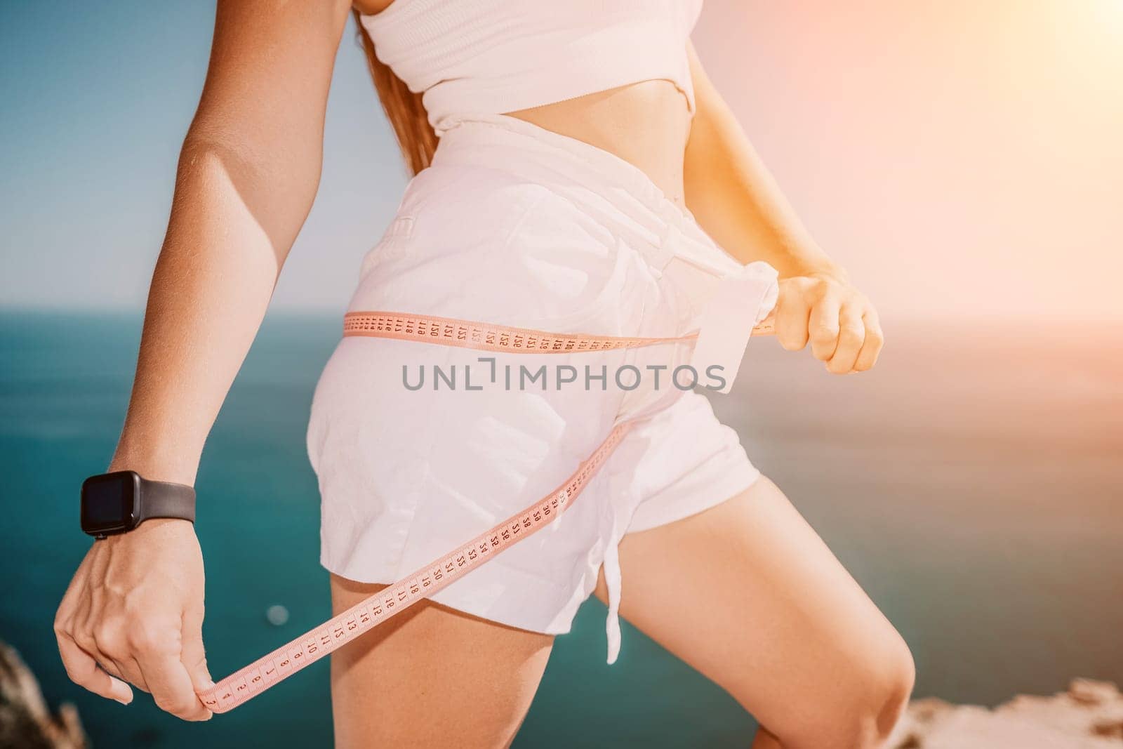 woman in white shorts holds in her hands pink measuring tape on her waist near the azure sea. Concept of detox and diet by panophotograph