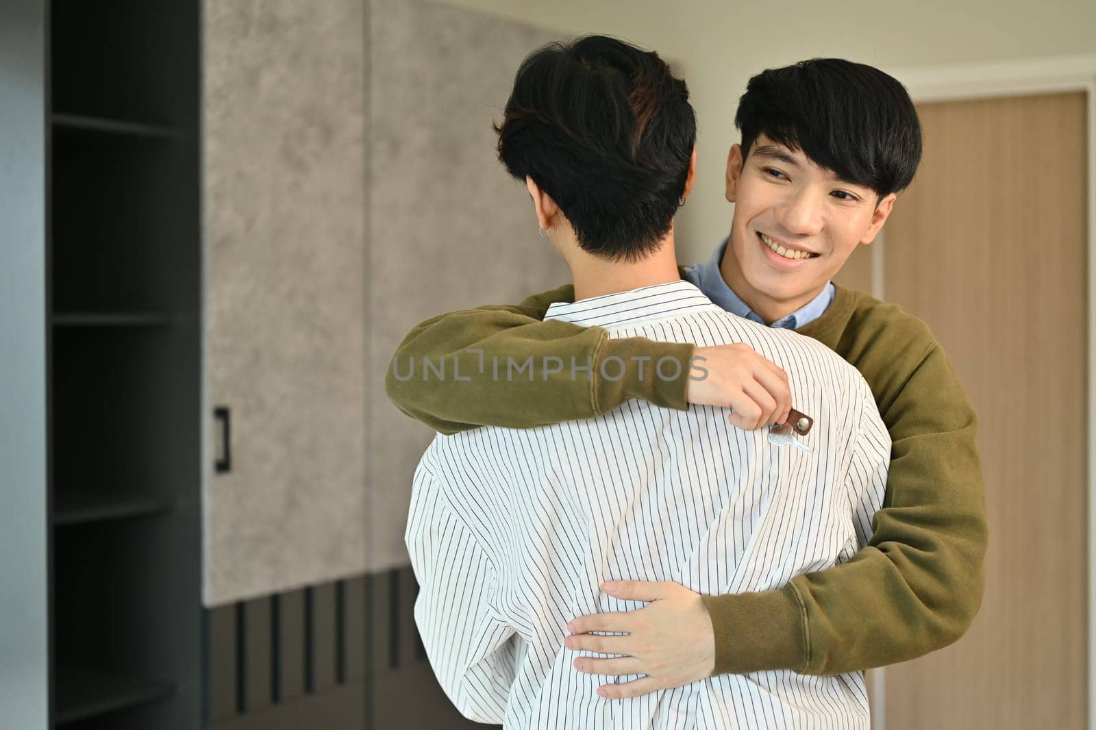 Happy smiling gay couple with keys celebrating moving into new home and hugging each other. LGBTQ, relocation and real estate property by prathanchorruangsak
