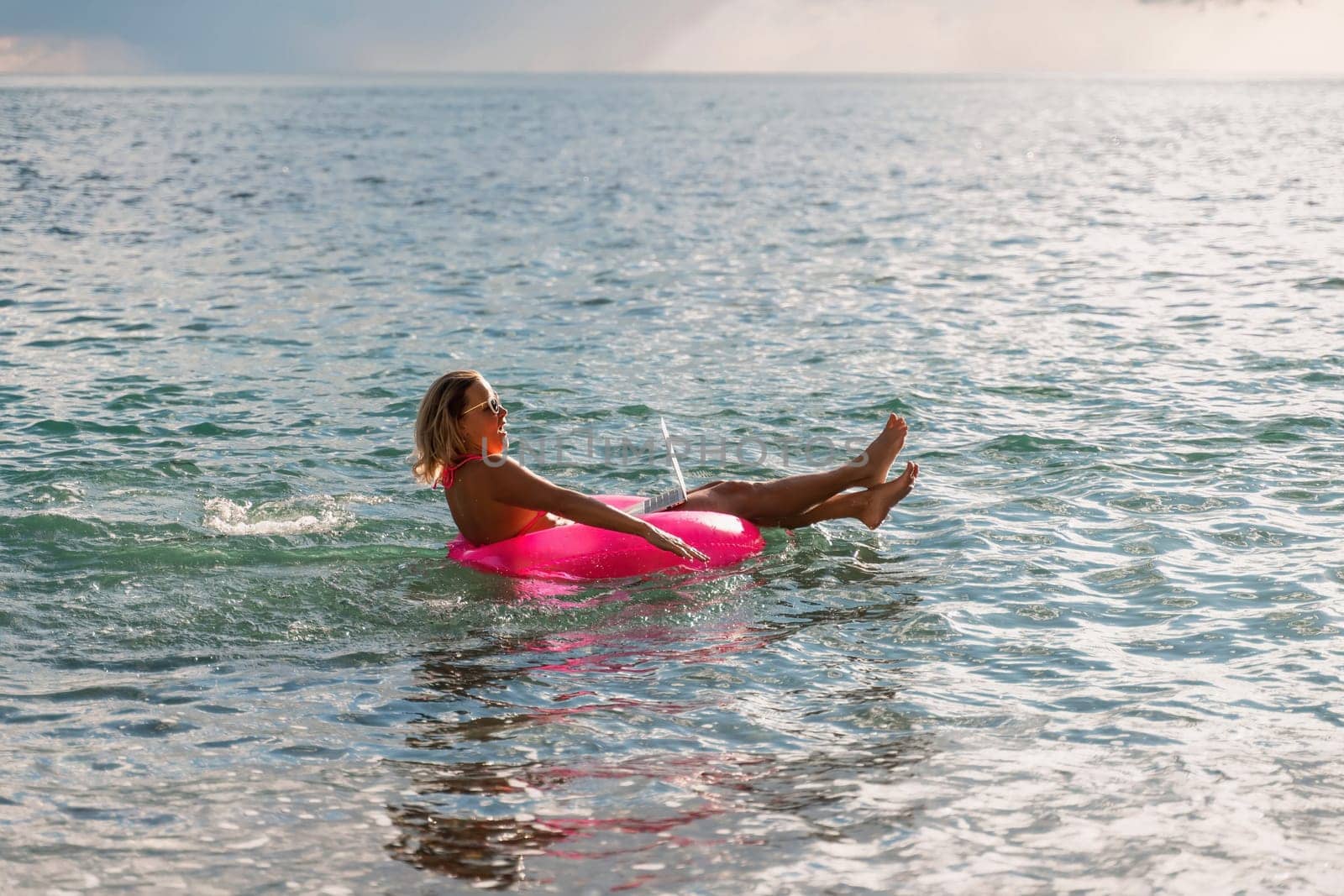 Woman works on laptop in sea. Freelancer, blond woman in sunglases floating on an inflatable big pink donut with a laptop in the sea at sunset. People summer vacation rest lifestyle concept