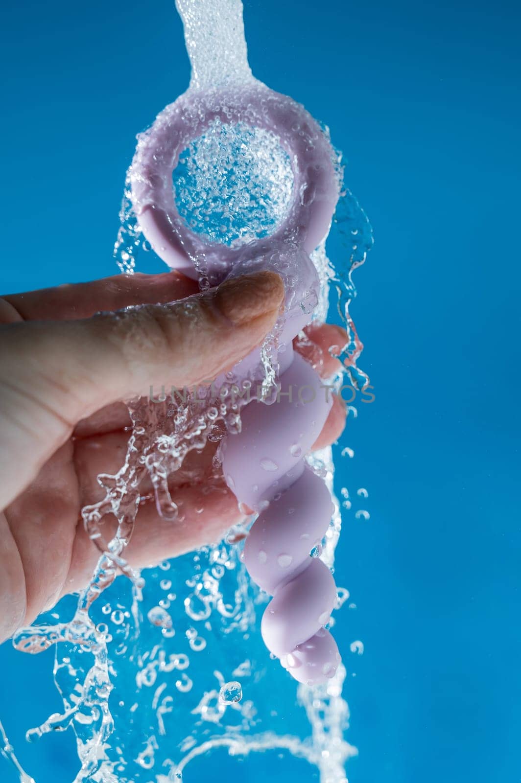 Woman holding lilac anal beads under running water on blue background. Sex toy hygiene concept