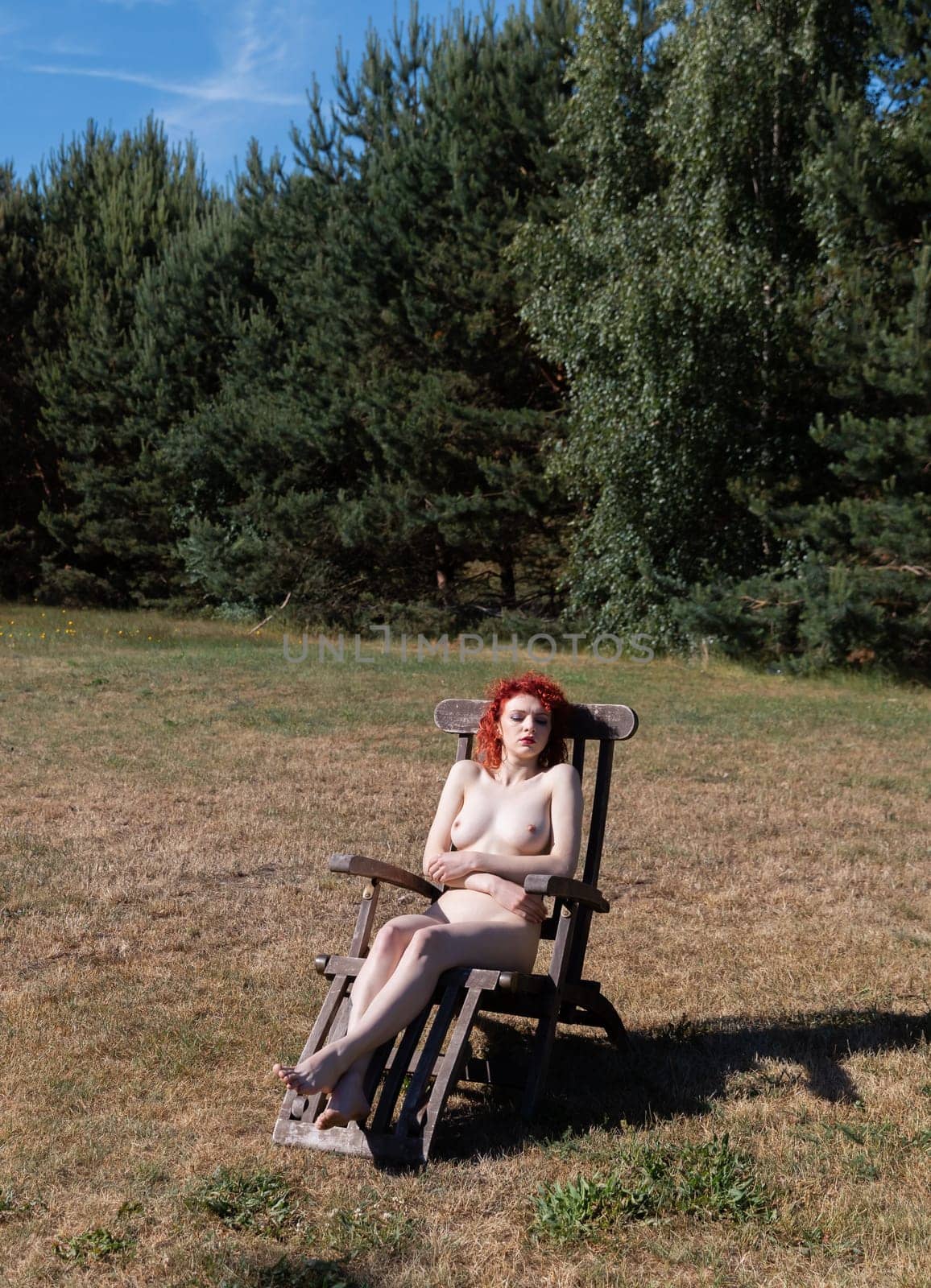 Young woman with scarlet hair poses nude sunbathing in a forest clearing sitting on a wooden country chair