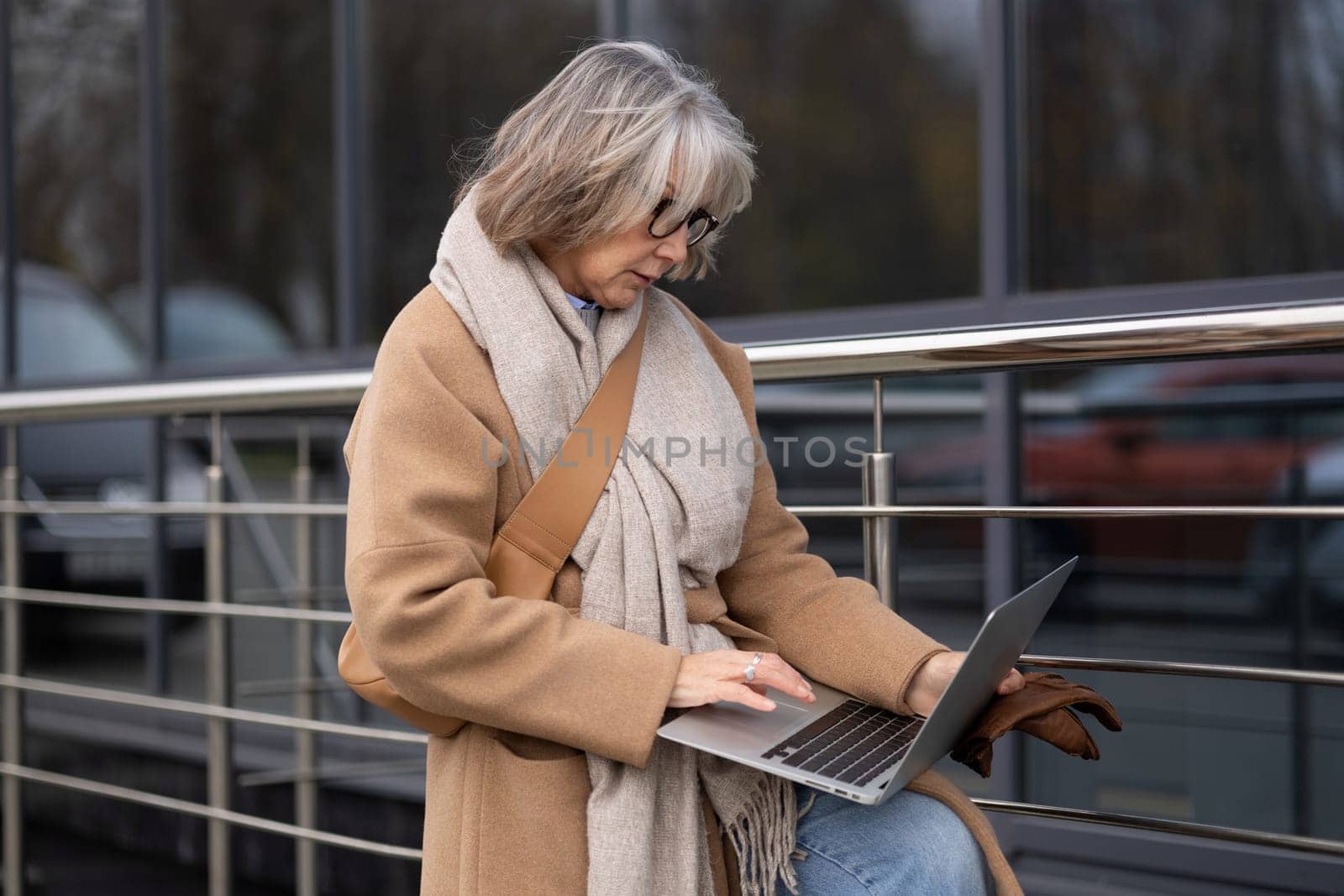 A mature woman, warmly dressed in a coat and scarf, focuses on her laptop while leaning against a railing, surrounded by a contemporary urban environment.