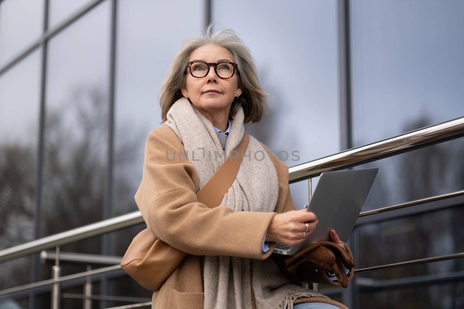 A senior woman, dressed warmly in a coat and scarf, sits on a railing outside a contemporary glass building, focused on her laptop as she enjoys her work in the chilly air.