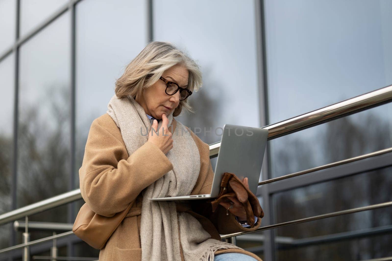 An older woman is focused on her laptop as she sits on a railing outdoors. She is dressed warmly in a coat and scarf, engaged in her work in a contemporary urban setting.