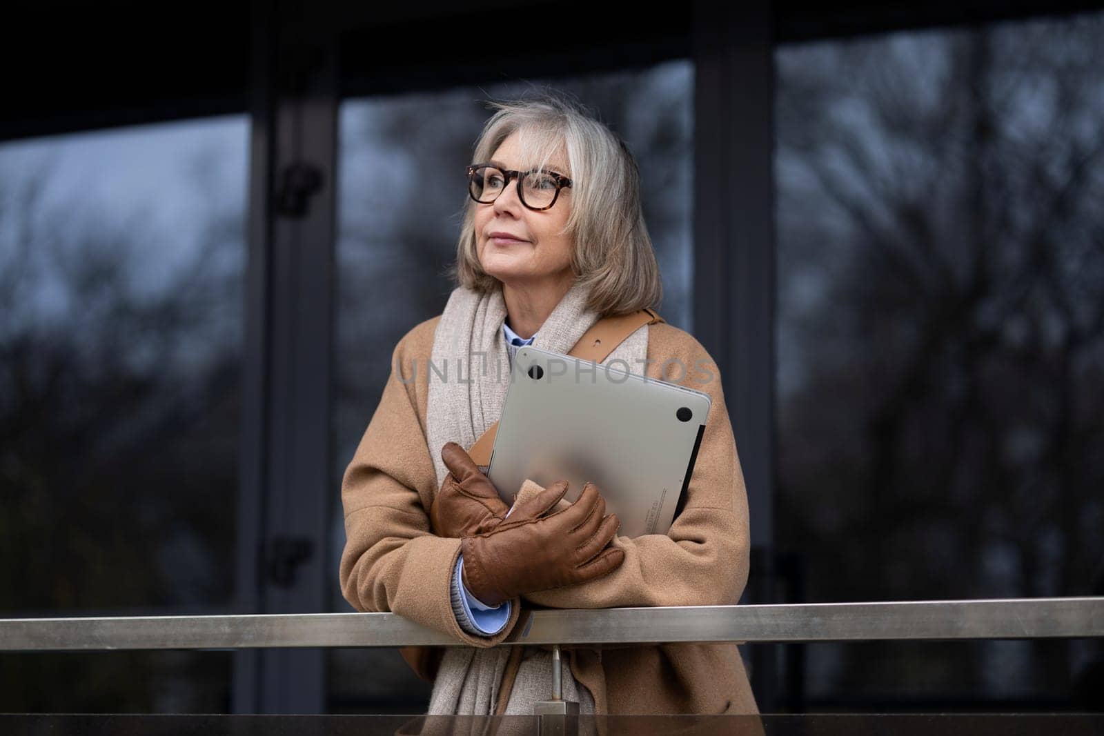 An elderly woman dressed warmly in a coat and gloves stands pensively by a railing. She holds a tablet close, gazing into the distance on a chilly autumn day in the city.