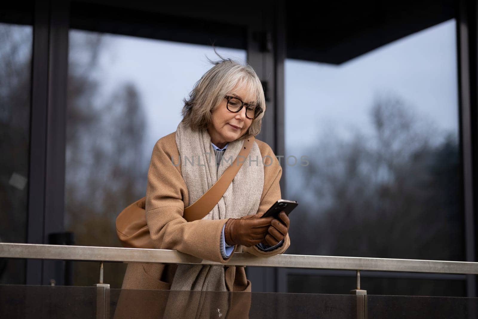 A mature woman stands on a balcony, focused on her smartphone. Dressed warmly, she enjoys the cool autumn air while checking her messages. The cloudy sky sets a tranquil mood.