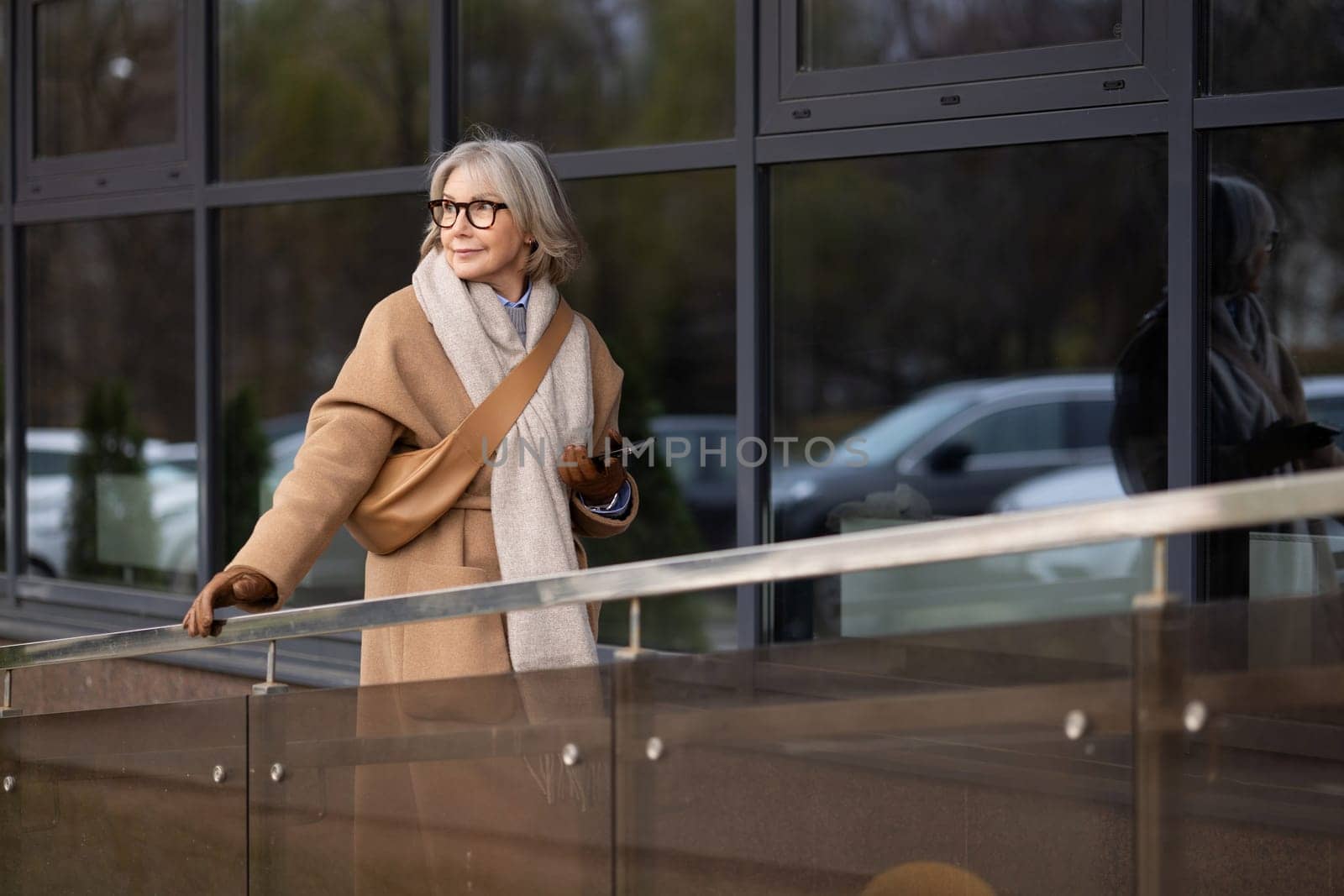 An elderly woman dressed in a fashionable coat and scarf stands beside a sleek glass building. She gazes thoughtfully while enjoying a sunny afternoon outdoors in a vibrant urban setting.