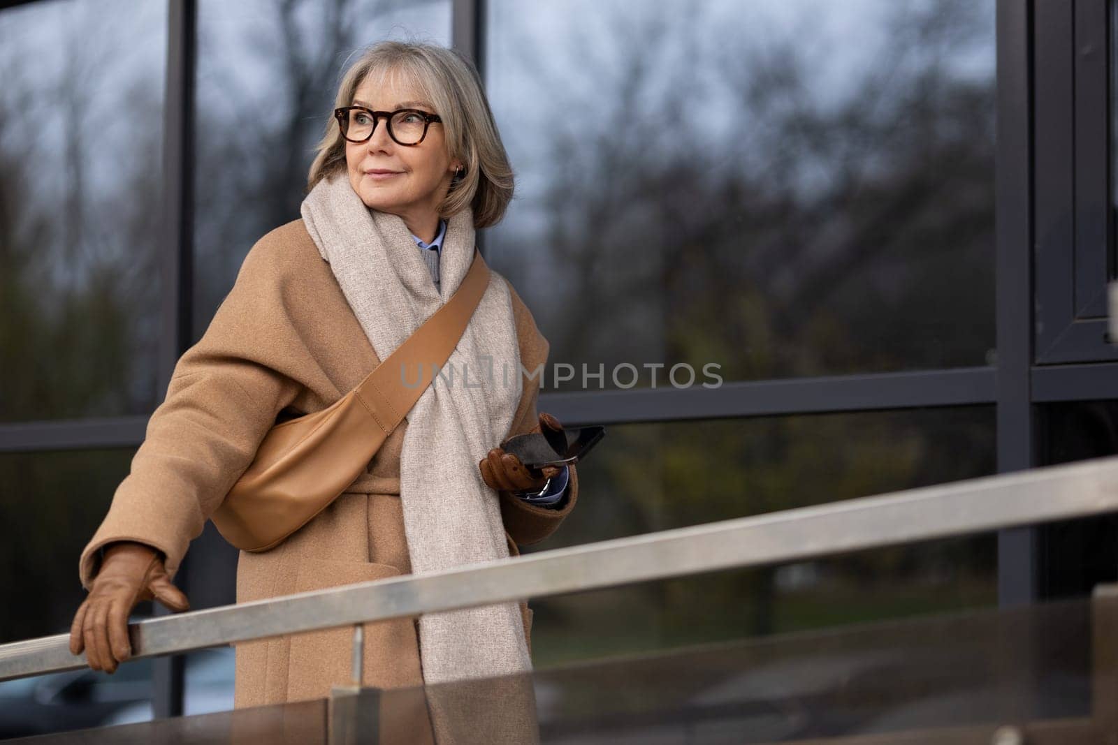 A fashionable woman stands near a railing, holding her phone and looking thoughtfully outside a contemporary building on a clear afternoon. Her outfit includes a stylish coat and scarf.
