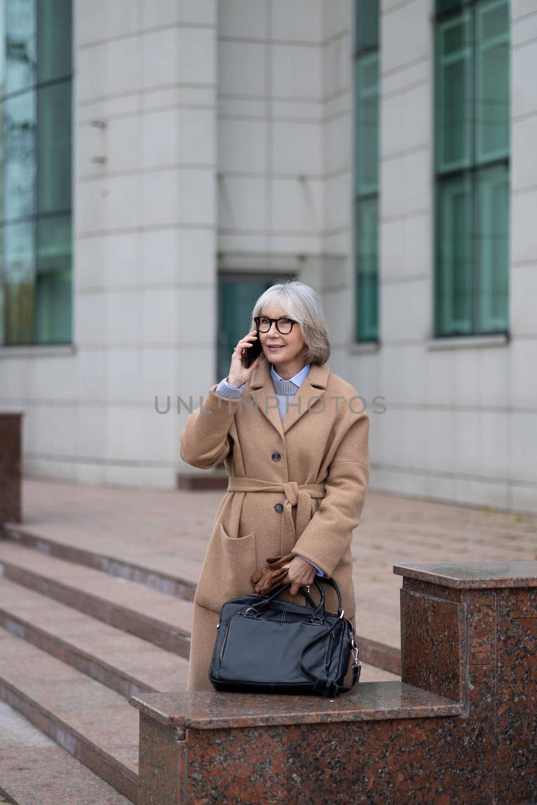 A mature woman with gray hair and glasses is engaged in a conversation on her phone. She stands confidently on stone steps, dressed in a fashionable coat, holding a handbag.