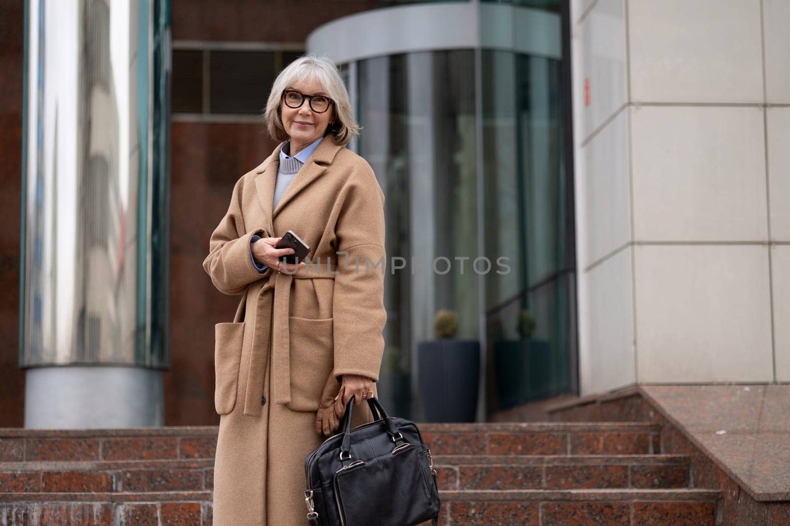 A woman dressed in a fashionable coat smiles while posing on the steps of a contemporary building, holding a smartphone and a black handbag under clear skies.