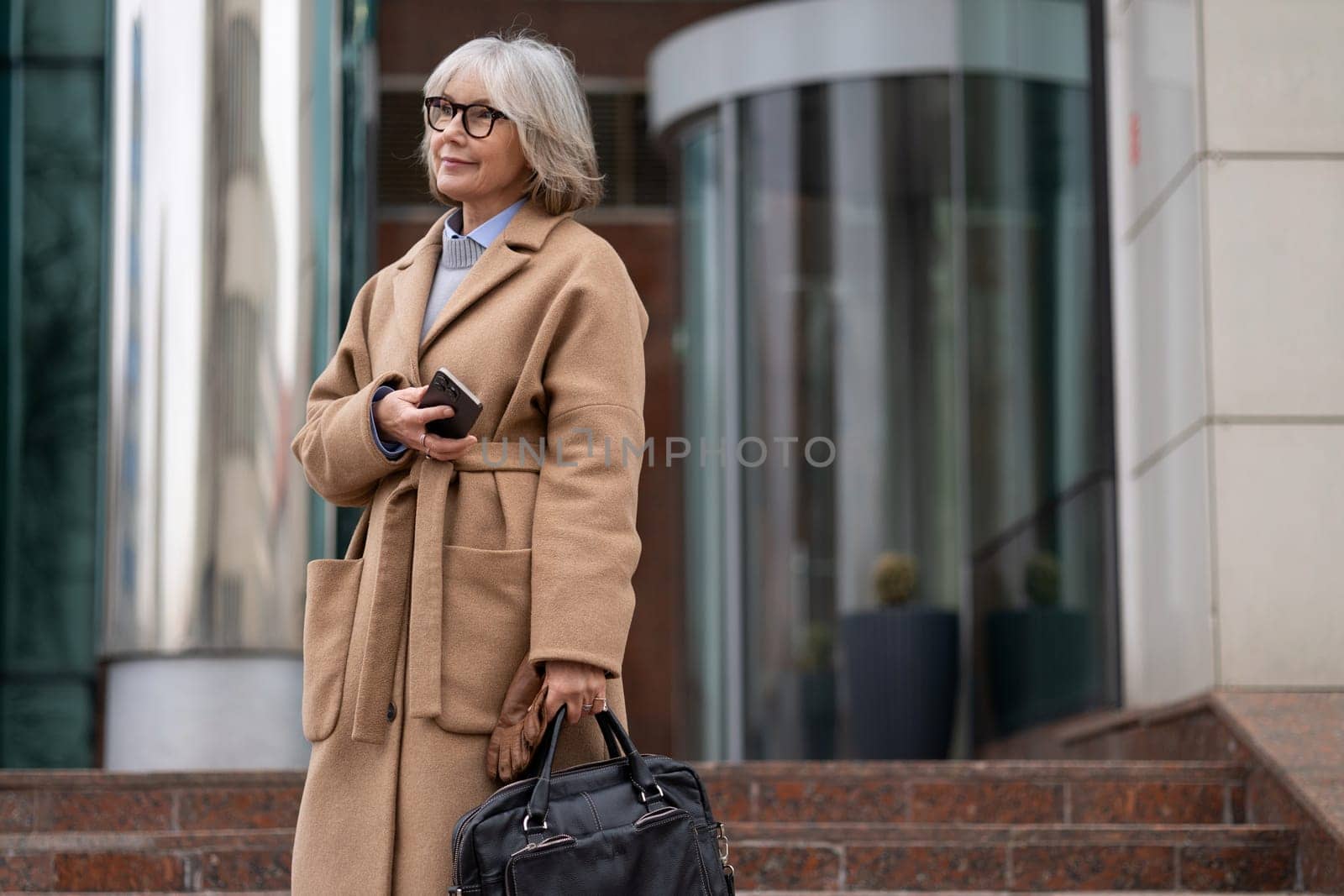 A mature woman with gray hair and glasses waits outside a contemporary building, dressed in a cozy coat, holding her phone and a handbag, exuding confidence.