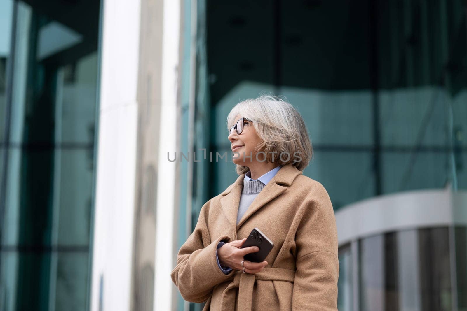 A senior woman with short gray hair, wearing glasses, stands outside a contemporary building, holding a phone and enjoying the daytime atmosphere in her stylish coat.