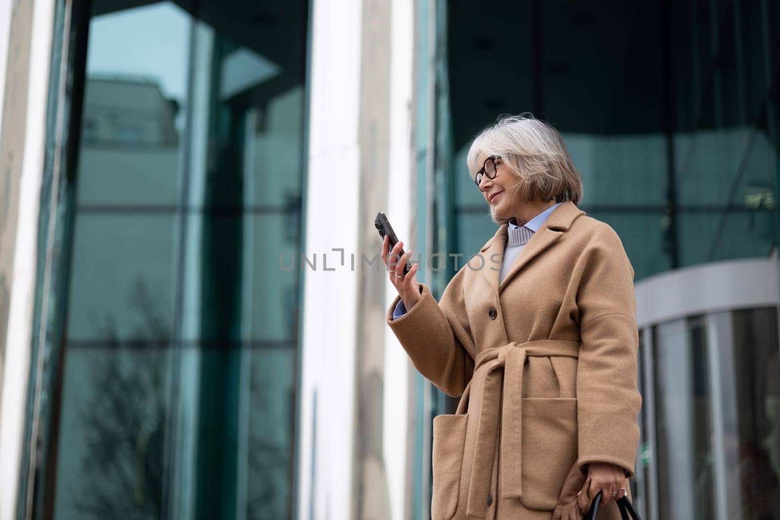 A senior woman with gray hair and glasses stands outside a sleek building while looking at her smartphone. She wears a fashionable beige coat and carries a bag, enjoying the day.