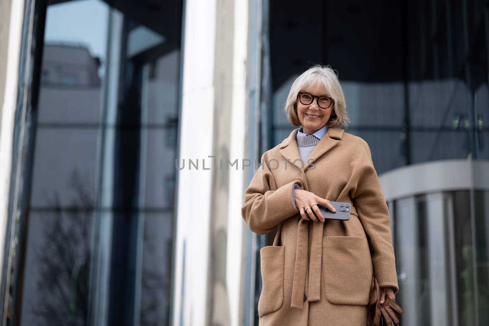 A smiling older woman stands outside a sleek modern building, wearing a fashionable coat and holding a smartphone. The scene captures a sunny day and an urban atmosphere.