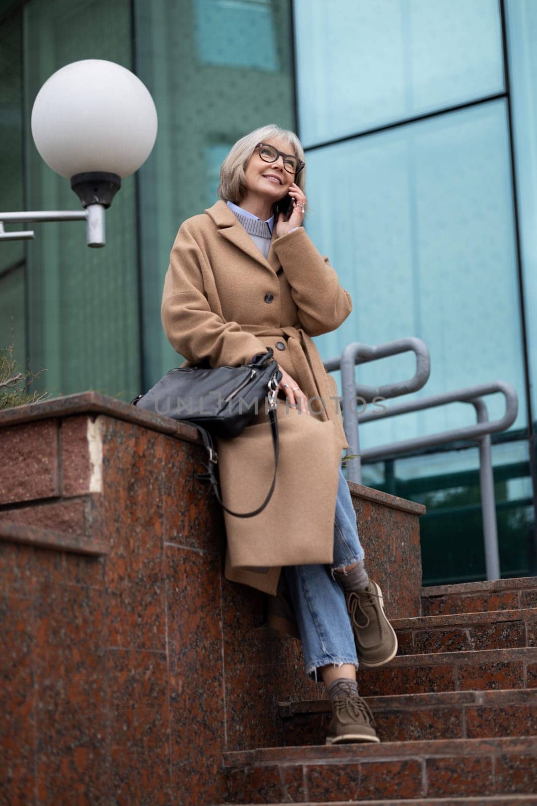 A smiling woman with glasses enjoys a conversation on her phone while seated on outdoor stairs. She carries a bag and is dressed casually in a fashionable coat, showcasing a relaxed vibe.