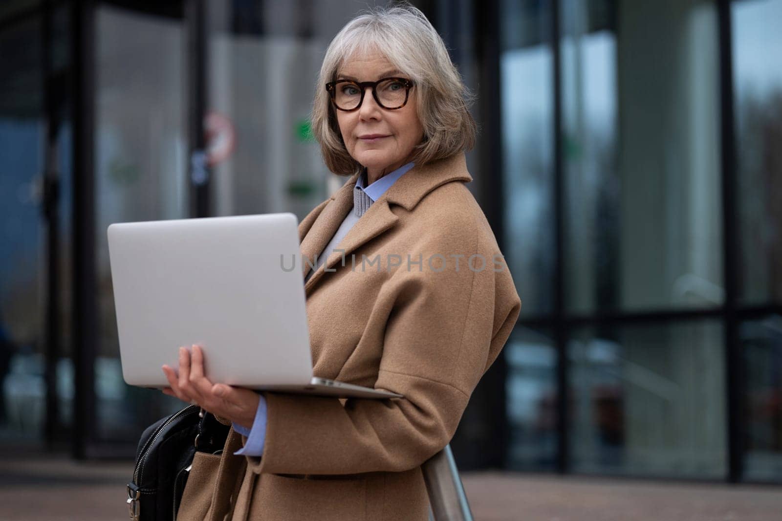 A senior woman stands confidently outside a contemporary building holding a laptop. Dressed in a stylish coat and glasses, she embodies professionalism in an urban environment.