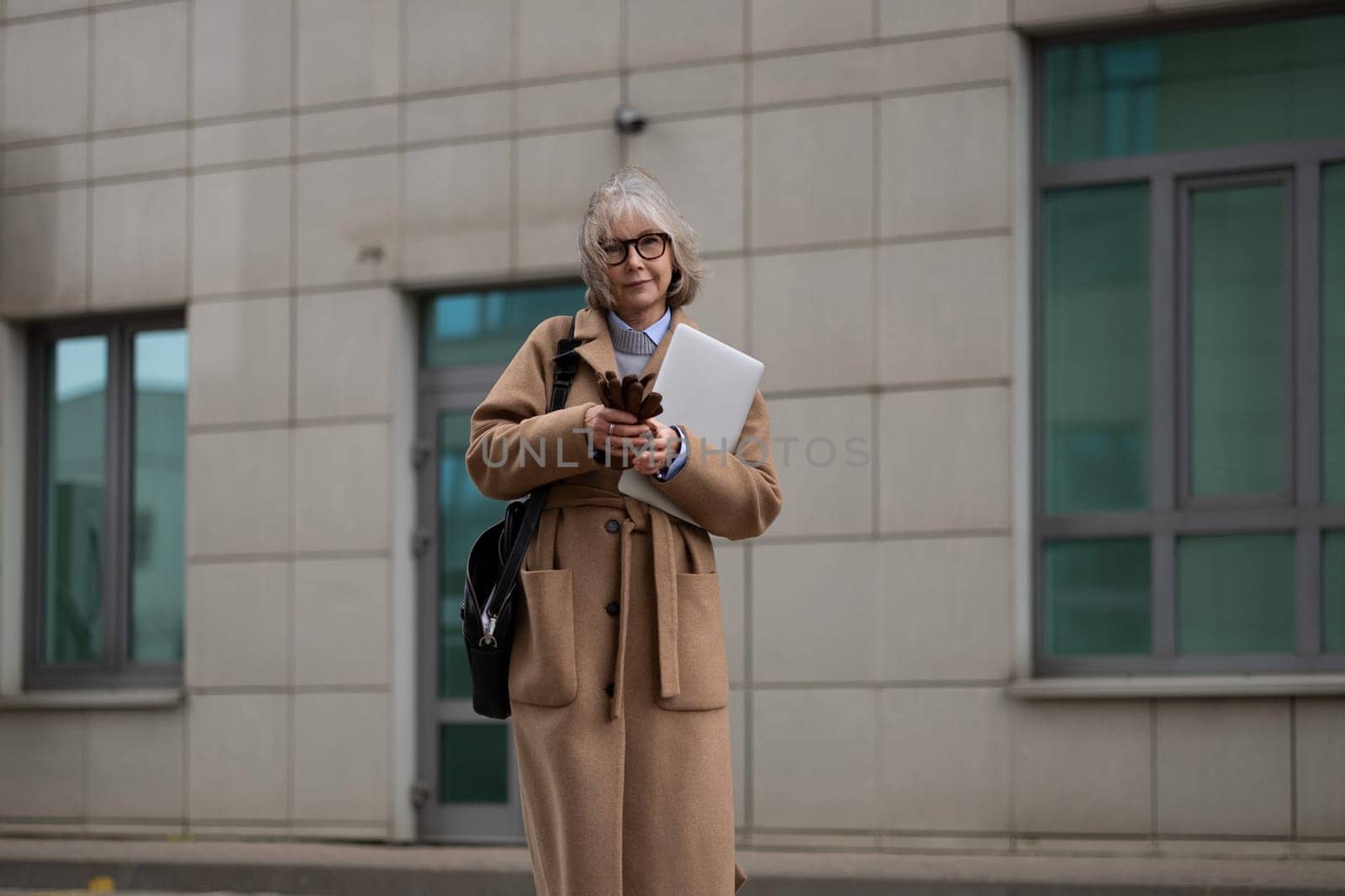 A business woman dressed in a beige coat stands outside a contemporary building while holding a laptop and checking her phone. The day appears overcast, adding a muted atmosphere.