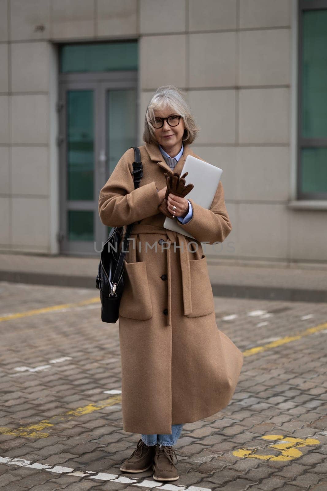 An older woman dressed in a fashionable long coat is standing outside a modern building. She holds a laptop and some notes, exuding confidence and style in a chilly environment.