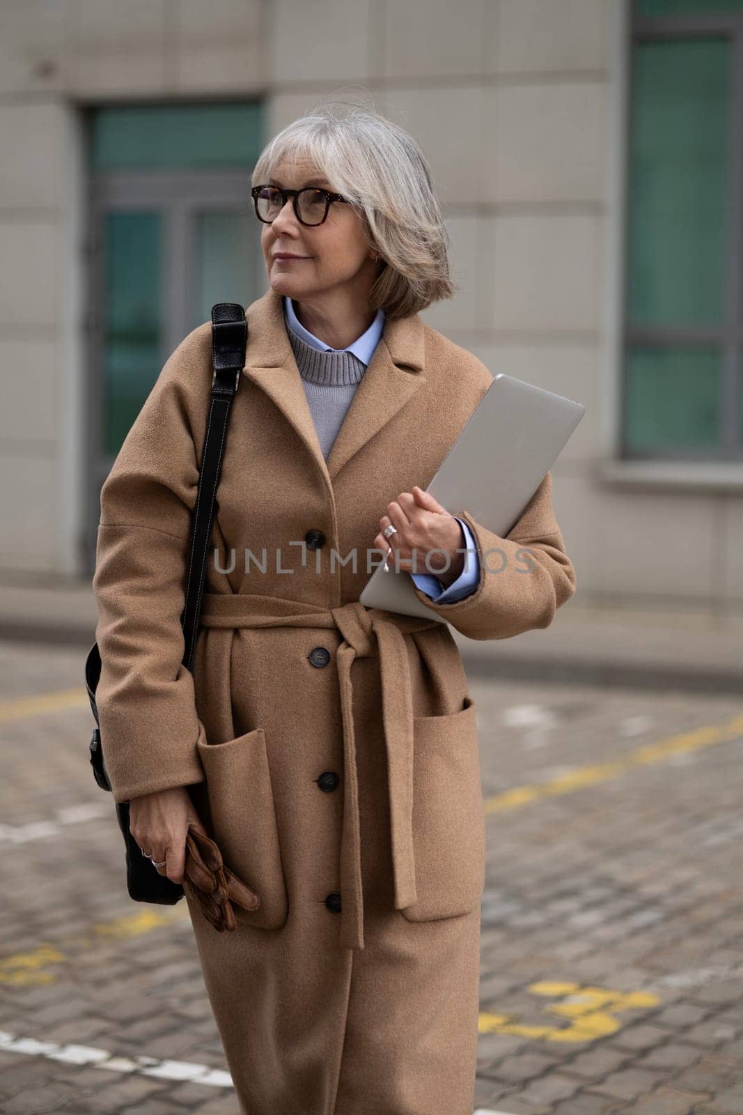 An older woman strolls down the street, wearing a chic coat and carrying a laptop. She exudes confidence and style amidst the modern urban surroundings.