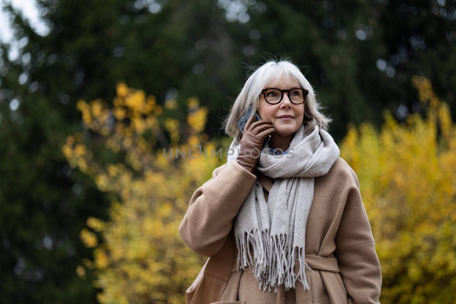 A senior woman stands in a park, wearing a stylish beige coat and a cozy scarf. She smiles while talking on her phone, showcasing the vibrant autumn foliage around her.